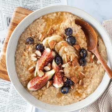 Ein cremiges Porridge mit frischem Obst und Mandeln auf einem hellen Untergrund, mit einer Zeitung und Kaffee im Bildrand.