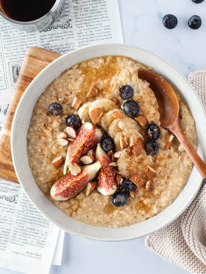 Ein cremiges Porridge mit frischem Obst und Mandeln auf einem hellen Untergrund, mit einer Zeitung und Kaffee im Bildrand.