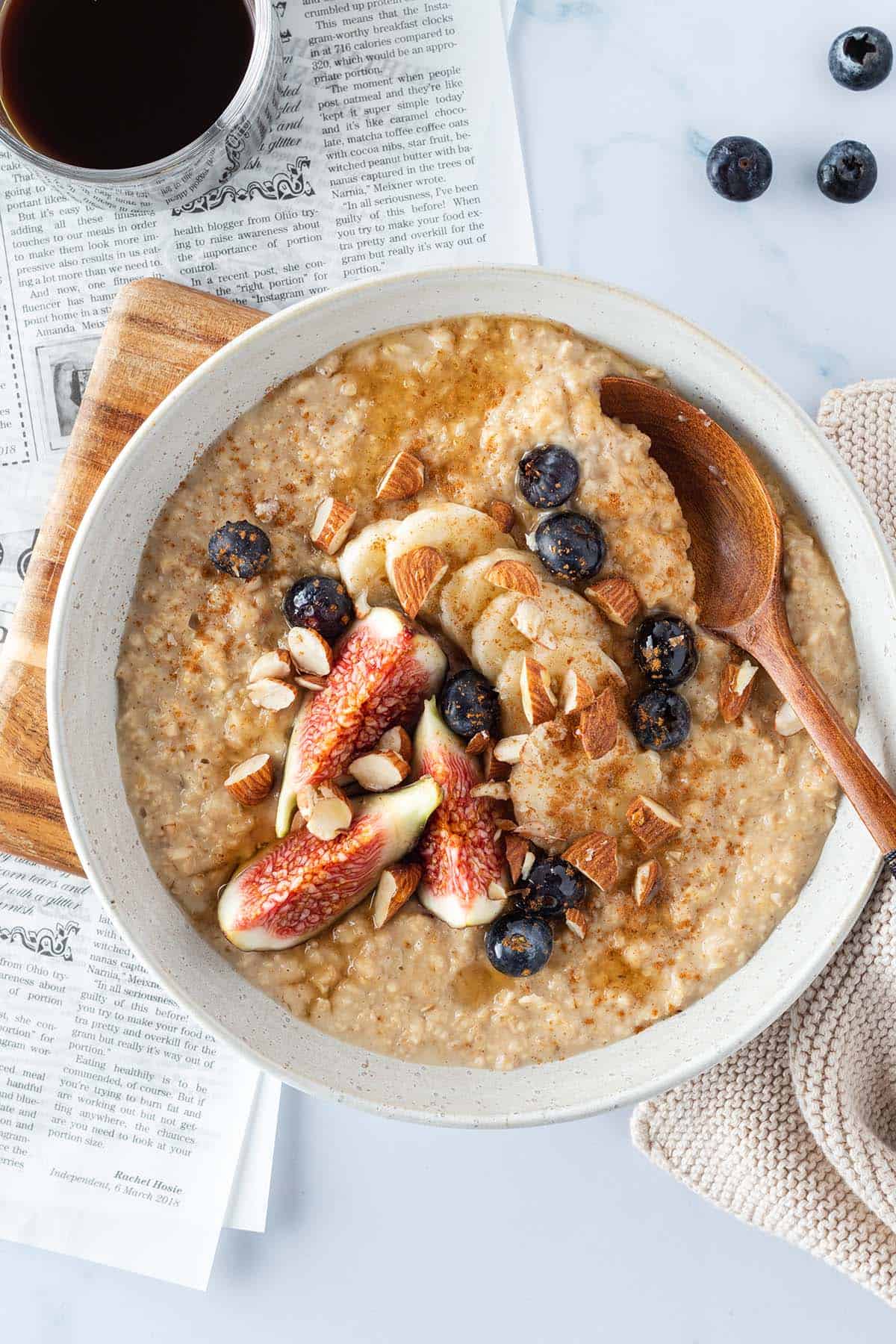 Ein cremiges Porridge mit frischem Obst und Mandeln auf einem hellen Untergrund, mit einer Zeitung und Kaffee im Bildrand.