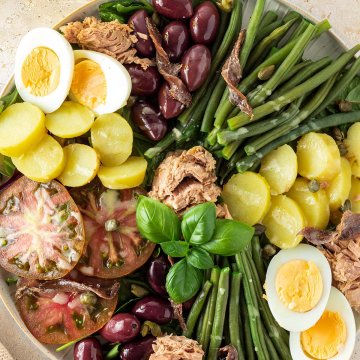 Bunter Nizzasalat serviert auf einem gro&szlig;en Teller mit Bohnen, Kartoffeln, Oliven, Sardellen, Thunfisch, Eiern, Tomaten und Kapern auf einem hellen Untergrund.