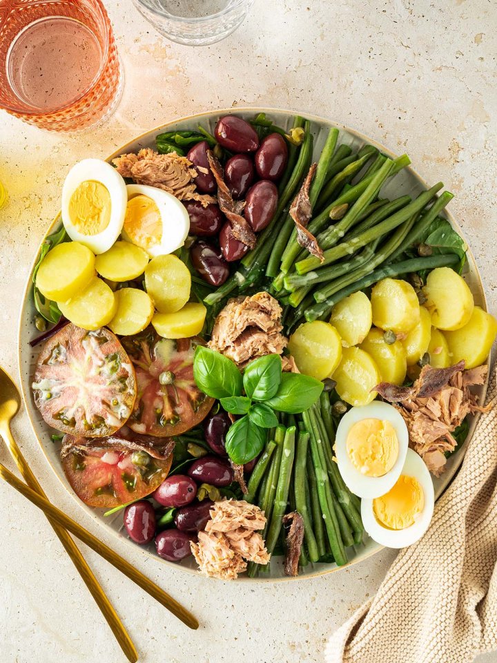 Nizza Salat mit Kartoffeln, Bohnen und Thunfisch auf einem beigen Untergrund mit mehreren Gl&auml;sern im Bildrand.