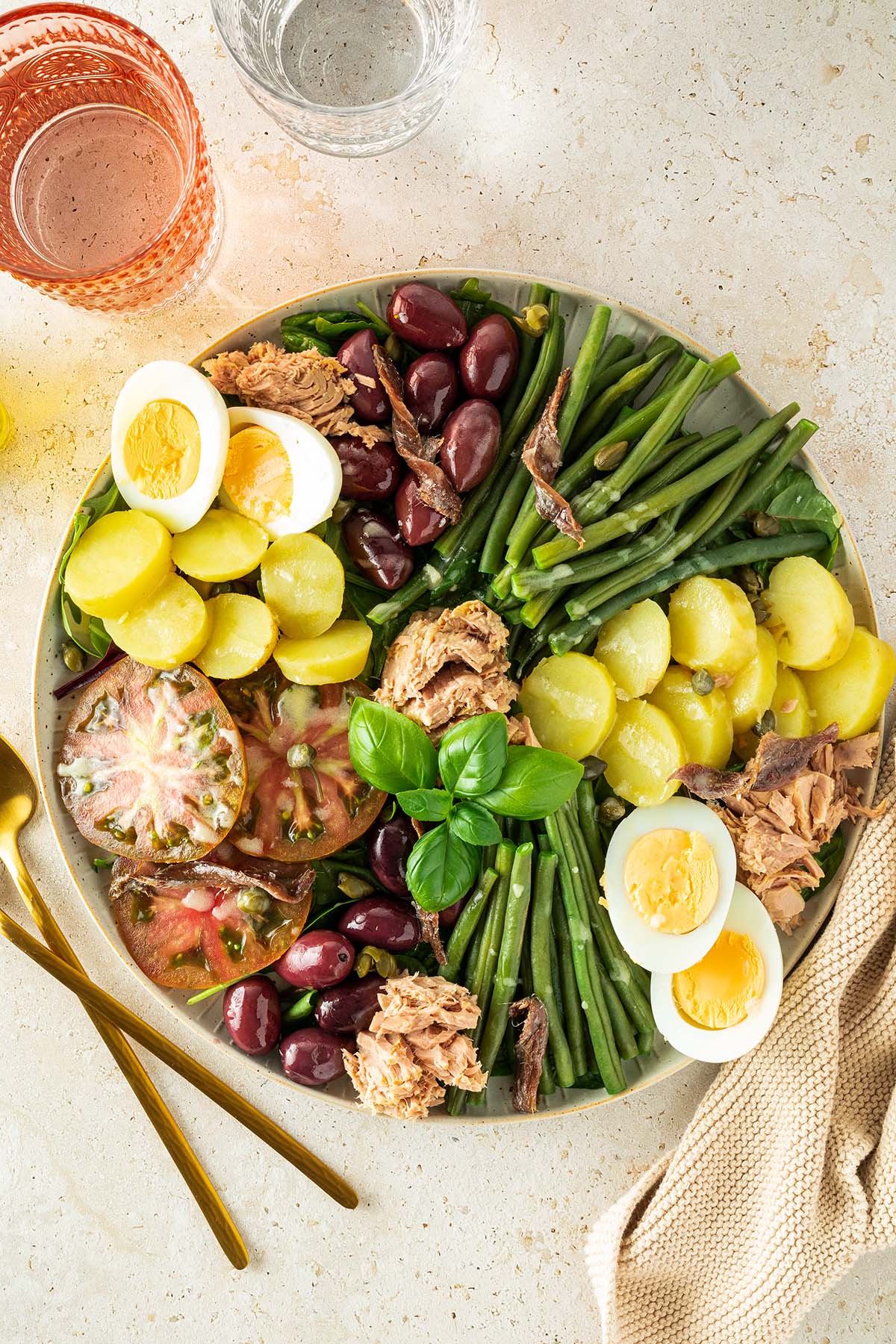 Nizza Salat mit Kartoffeln, Bohnen und Thunfisch auf einem beigen Untergrund mit mehreren Gl&auml;sern im Bildrand.
