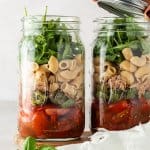 Zwei Portionen Nudelsalat im Glas stehen nebeneinander vor einem beigen Hintergrund, wobei von einem der beiden Gl&auml;ser gerade der Deckel abgenommen wird.