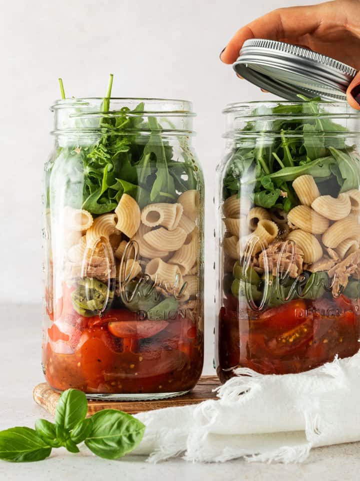 Zwei Portionen Nudelsalat im Glas stehen nebeneinander vor einem beigen Hintergrund, wobei von einem der beiden Gl&auml;ser gerade der Deckel abgenommen wird.