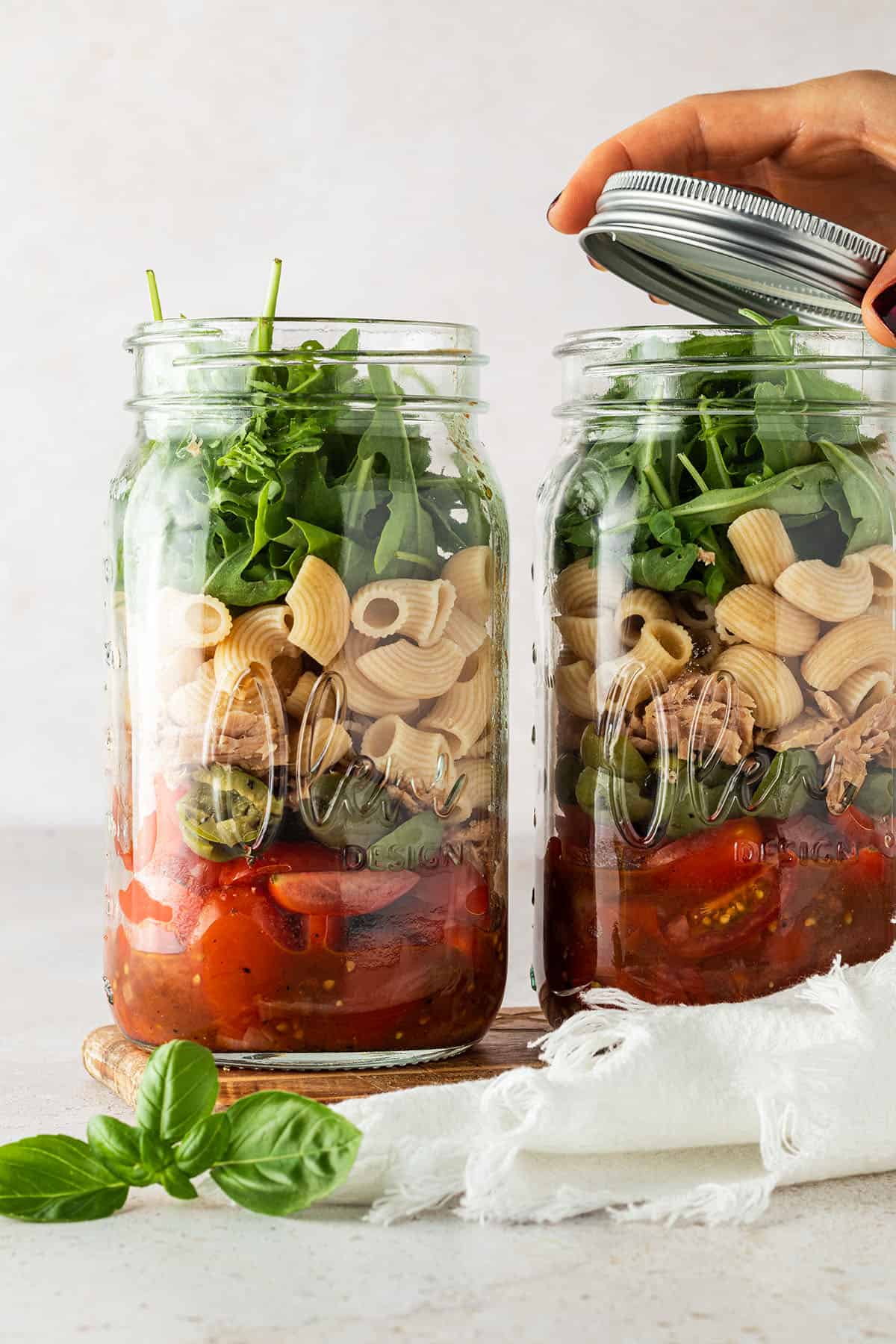 Zwei Portionen Nudelsalat im Glas stehen nebeneinander vor einem beigen Hintergrund, wobei von einem der beiden Gl&auml;ser gerade der Deckel abgenommen wird.