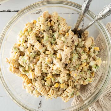 Ein cremiger Nudelsalat mit Thunfisch, Mais und Gemüse in einer großen Schüssel, Teller und ein Tuch im Bildrand.