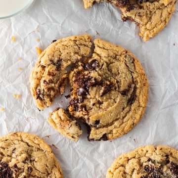 Vier gro&szlig;e Chocolate Chip Cookies auf Backpapier, einer angebissen, einer angebrochen mit sichtbar geschmolzener Schokolade, daneben ein Glas Milch.