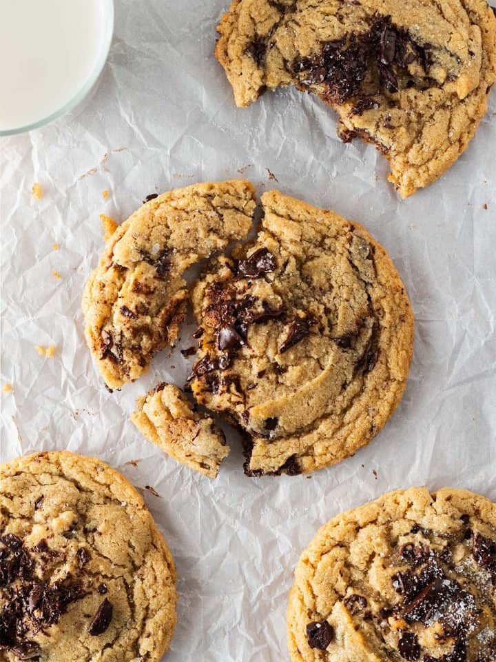 Vier gro&szlig;e Chocolate Chip Cookies auf Backpapier, einer angebissen, einer angebrochen mit sichtbar geschmolzener Schokolade, daneben ein Glas Milch.