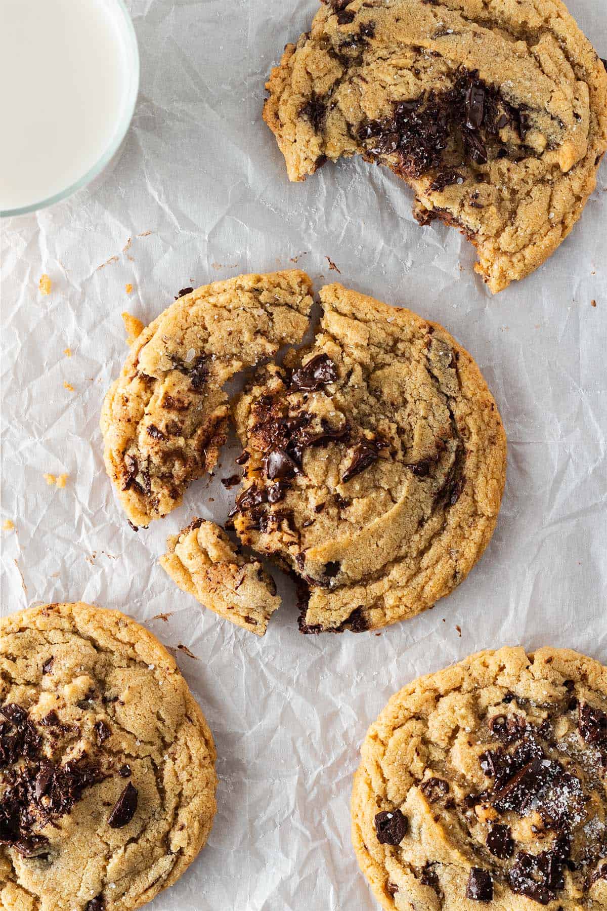Vier gro&szlig;e Chocolate Chip Cookies auf Backpapier, einer angebissen, einer angebrochen mit sichtbar geschmolzener Schokolade, daneben ein Glas Milch.