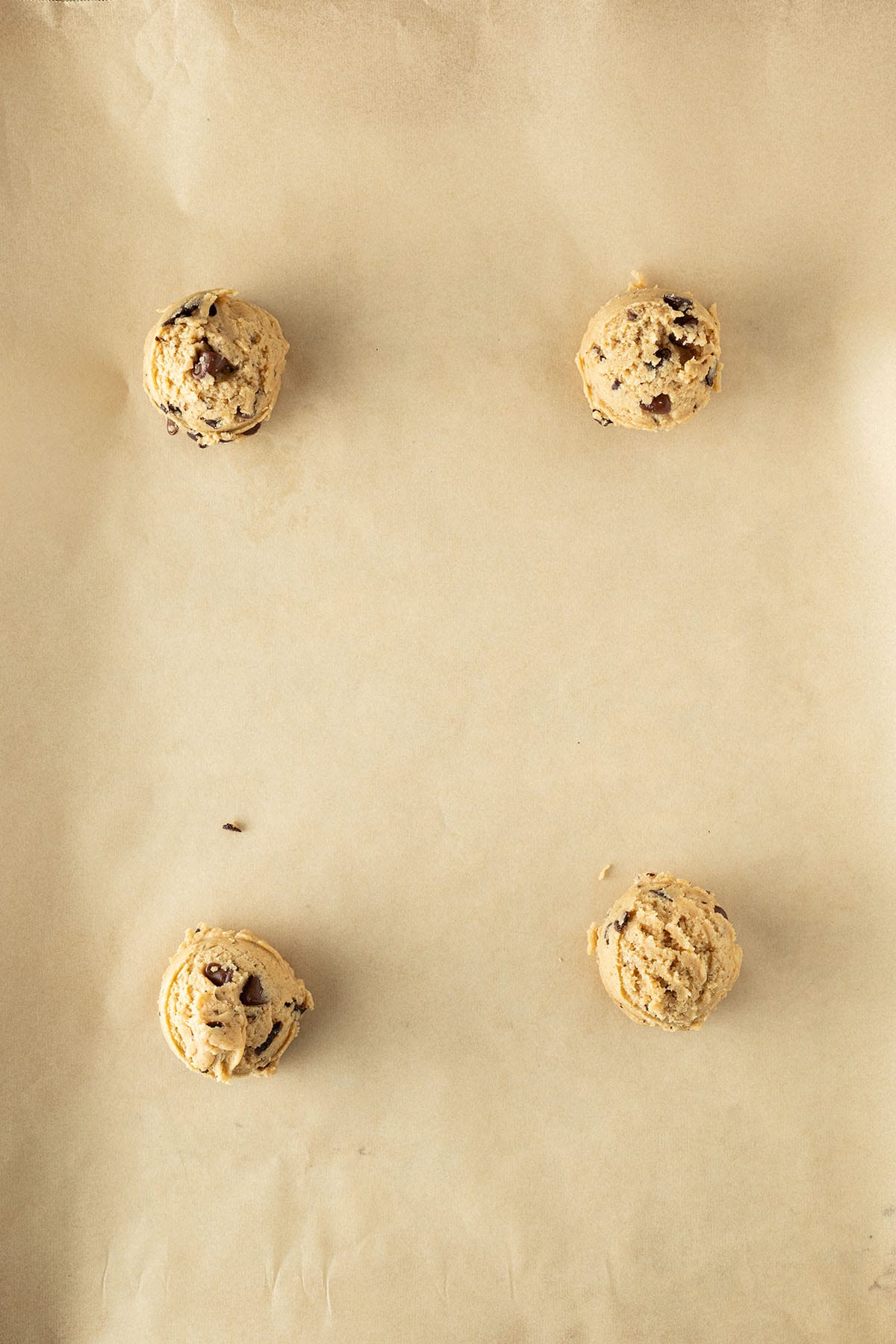 Vier Kugeln Chocolate-Chip-Cookie-Teig gleichm&auml;&szlig;ig auf Backpapier verteilt, vor dem Backen.