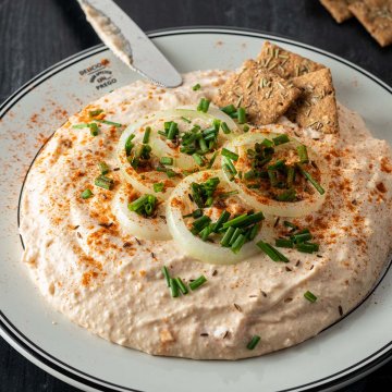 Cremiger Obazda serviert mit Zwiebelringen, Schnittlauch und Cracker auf einem dunklen Holzuntergrund.
