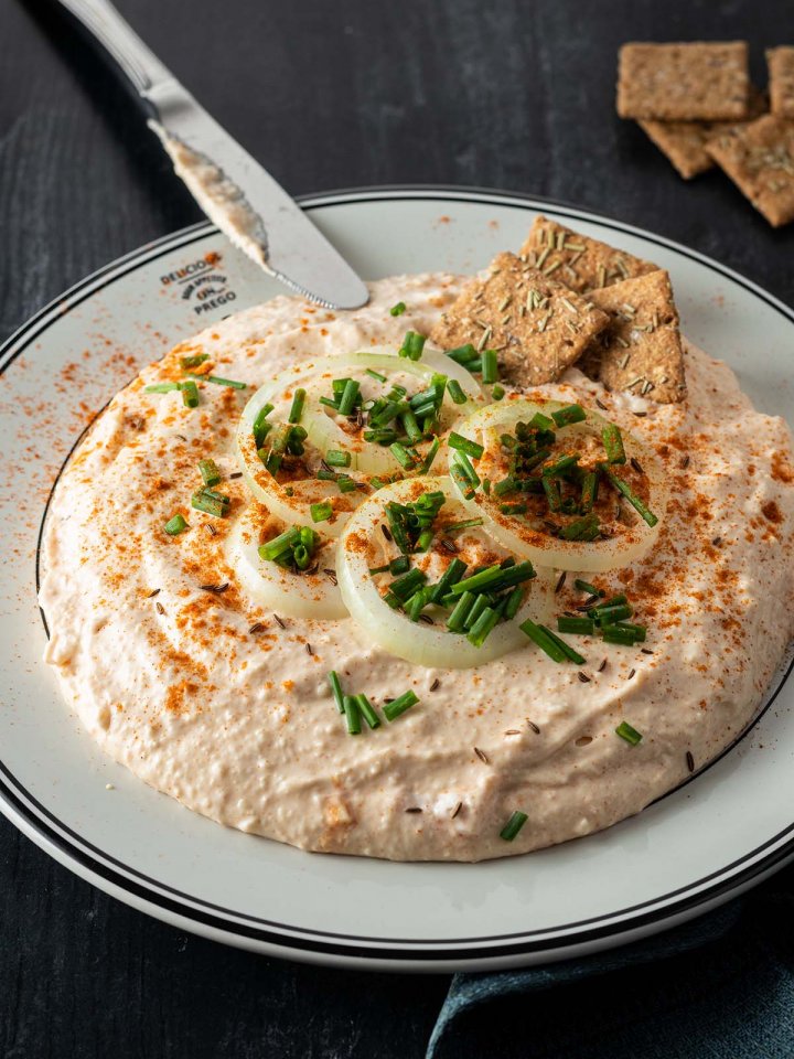 Cremiger Obazda serviert mit Zwiebelringen, Schnittlauch und Cracker auf einem dunklen Holzuntergrund.