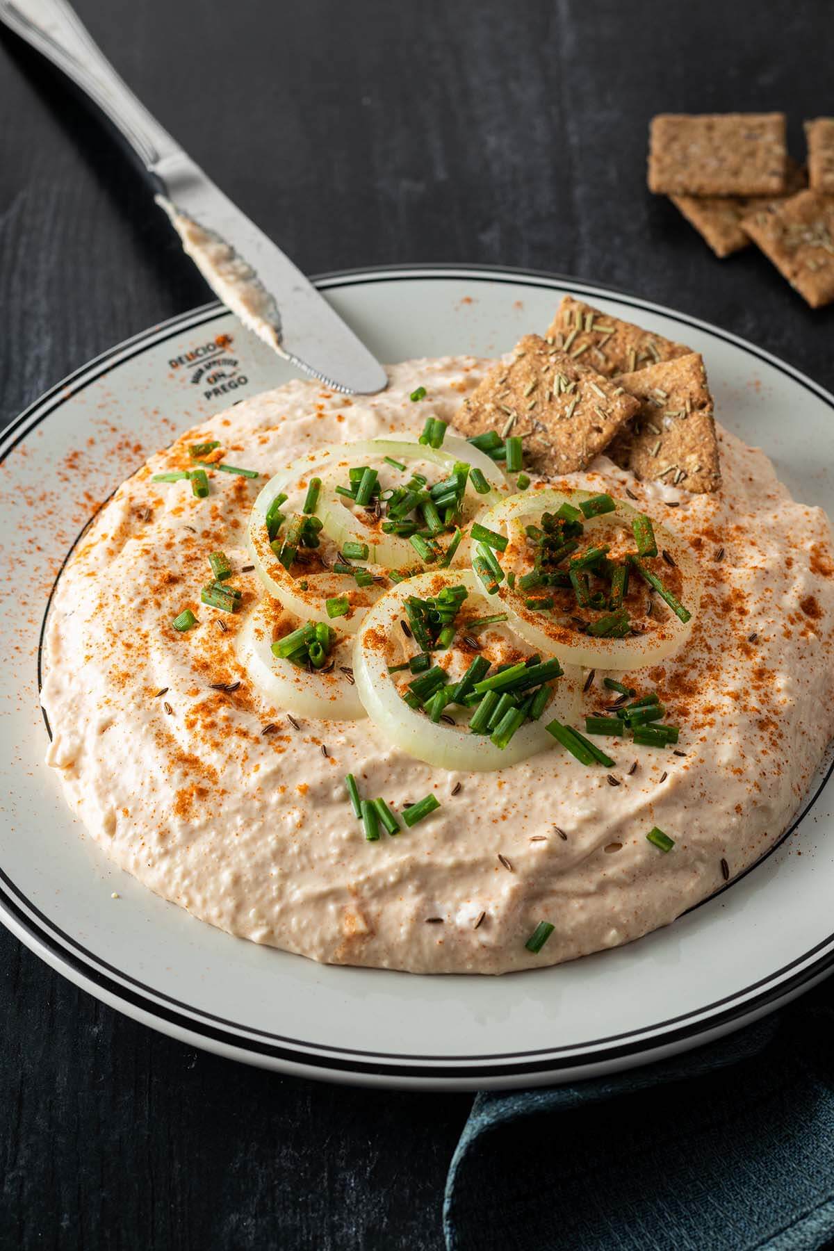 Cremiger Obazda serviert mit Zwiebelringen, Schnittlauch und Cracker auf einem dunklen Holzuntergrund.