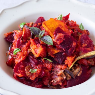 Farbenfrohes Ofen K&uuml;rbisgem&uuml;se mit Butternutk&uuml;rbis, Aubergine und Roter Bete, das mit Pesto verfeinert wurde, serviert in einem wei&szlig;en Teller auf einem wei&szlig;en Holztisch.