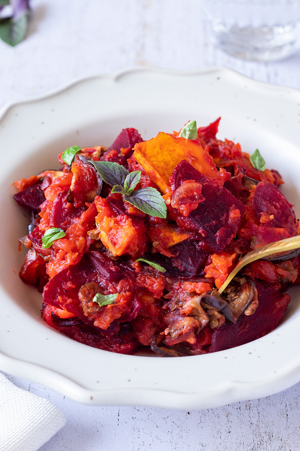 Farbenfrohes Ofen K&uuml;rbisgem&uuml;se mit Butternutk&uuml;rbis, Aubergine und Roter Bete, das mit Pesto verfeinert wurde, serviert in einem wei&szlig;en Teller auf einem wei&szlig;en Holztisch.