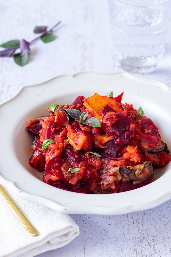 Farbenfrohes Ofen K&uuml;rbisgem&uuml;se mit Butternutk&uuml;rbis, Aubergine und Roter Bete, das mit Pesto verfeinert wurde, serviert in einem wei&szlig;en Teller auf einem wei&szlig;en Holztisch.