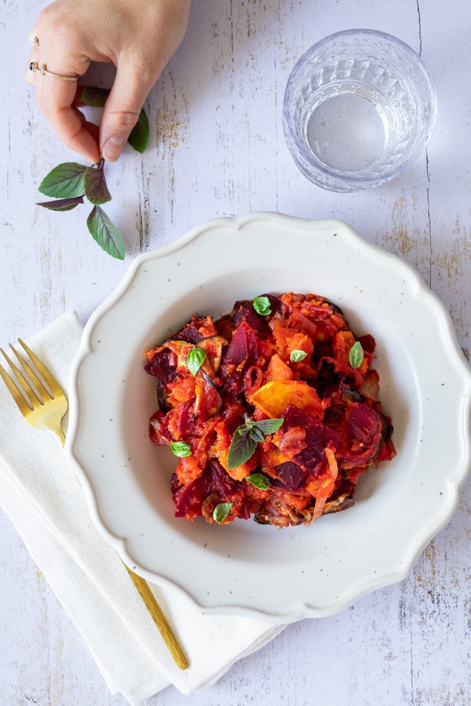 Farbenfrohes Ofen K&uuml;rbisgem&uuml;se mit Butternutk&uuml;rbis, Aubergine und Roter Bete, das mit Pesto verfeinert wurde, serviert in einem wei&szlig;en Teller auf einem wei&szlig;en Holztisch.