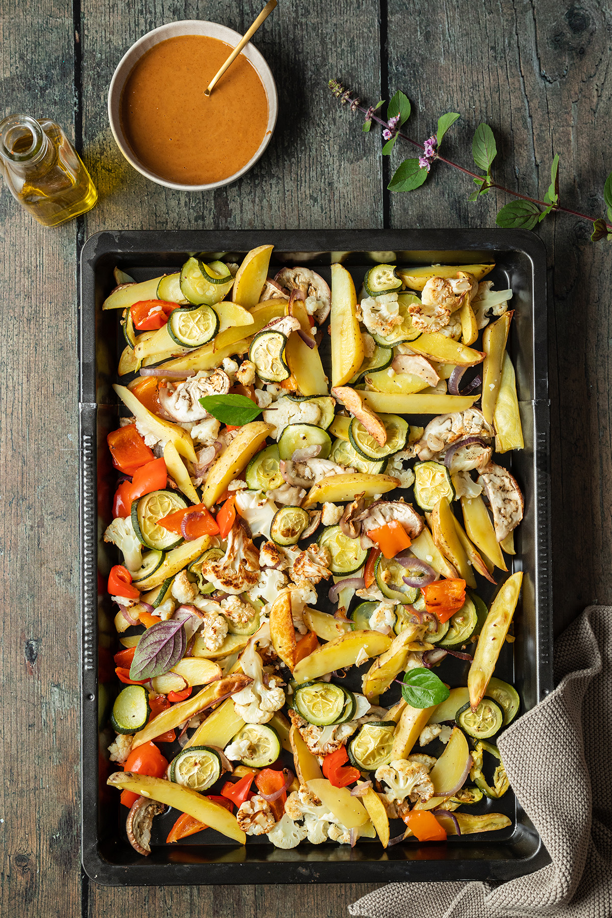 Blech mit Ofengem&uuml;se mit Kartoffel Spalten auf dunklem Holztisch, daneben eine Sch&uuml;ssel mit Erdnusssauce und eine Flasche Oliven&ouml;l.
