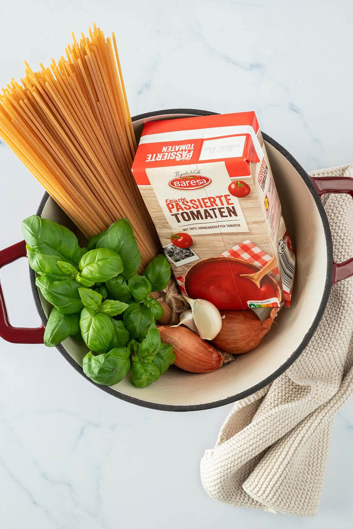 Topf mit ungekochten Spaghetti, einer Packung passierter Tomaten, Knoblauchzehen, Schalotten und frischem Basilikum.