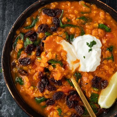 Nahaufnahme von einer Sch&uuml;ssel mit einer orientalischen, roten Linsensuppe, die mit Joghurt, Rosinen, Limette und frischen Kr&auml;utern serviert wird.