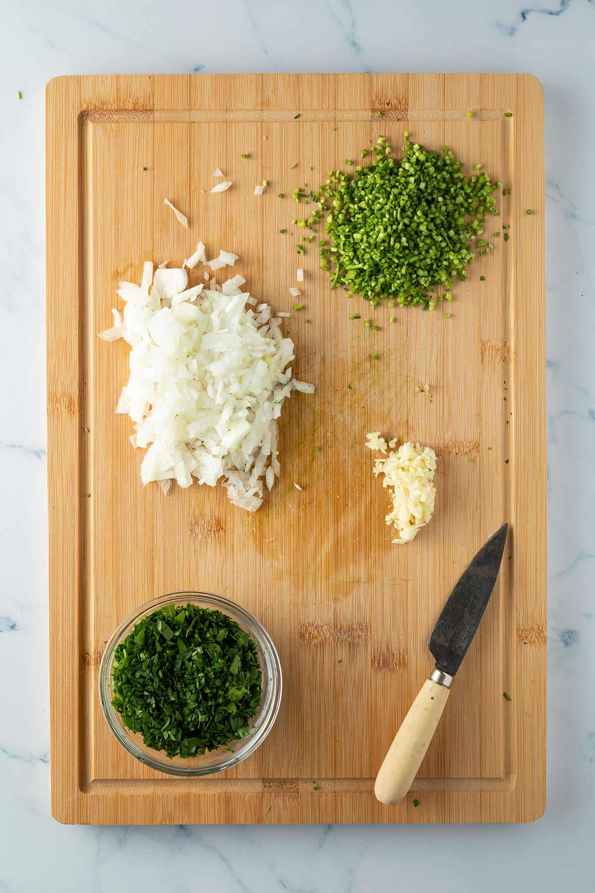 Fein gew&uuml;rfelte Zwiebeln, Knoblauch, Petersilienst&auml;ngel und -Bl&auml;tter auf einem Brett.
