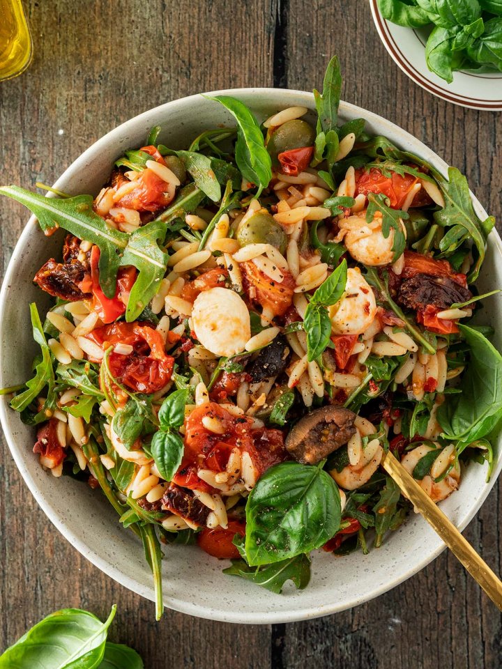 Vogelperspektive auf einen Teller mit einem Kritharaki-Nudelsalat mit Tomaten, Oliven, Mozzarella, Rucola und Tomaten.