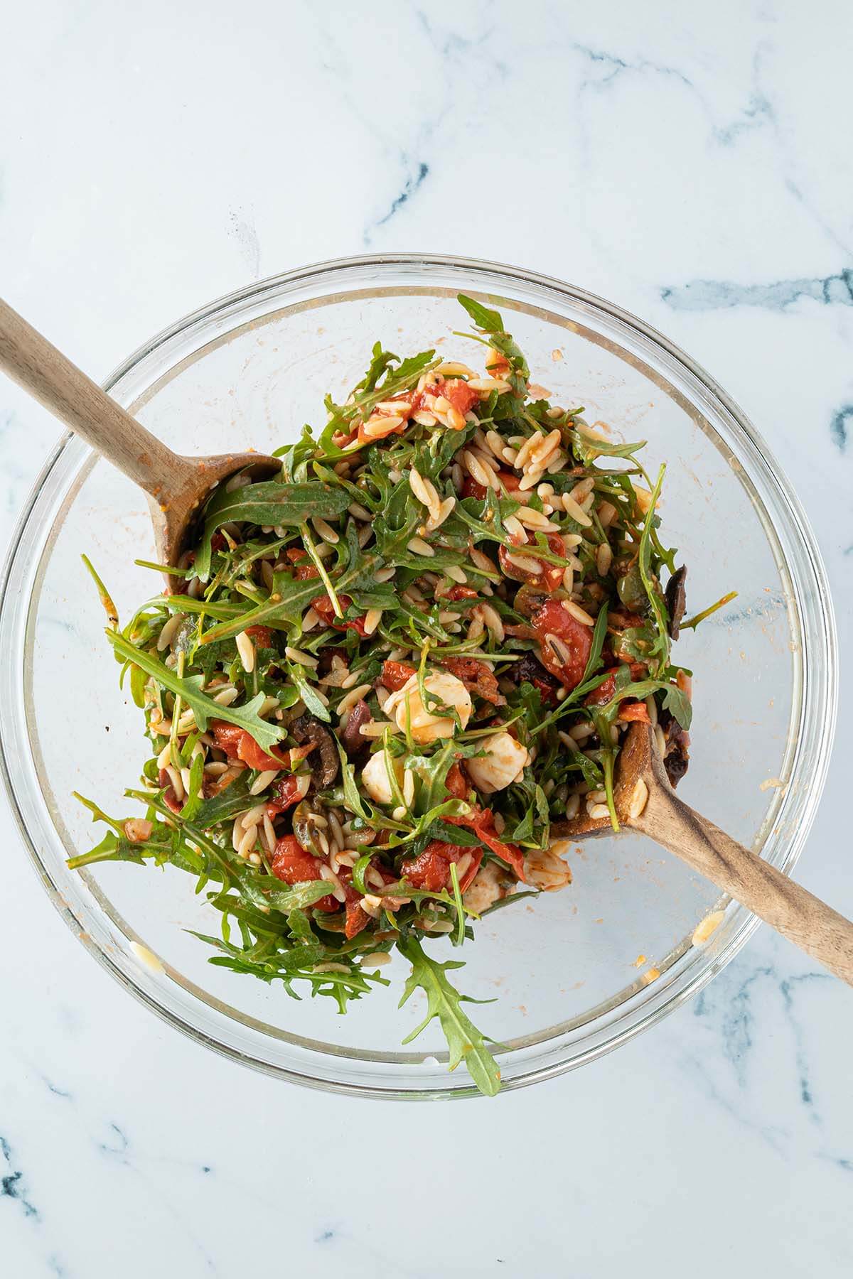Zutaten f&uuml;r den Orzo Salat werden in einer gro&szlig;en Glassch&uuml;ssel mit Salatbesteck vermischt.