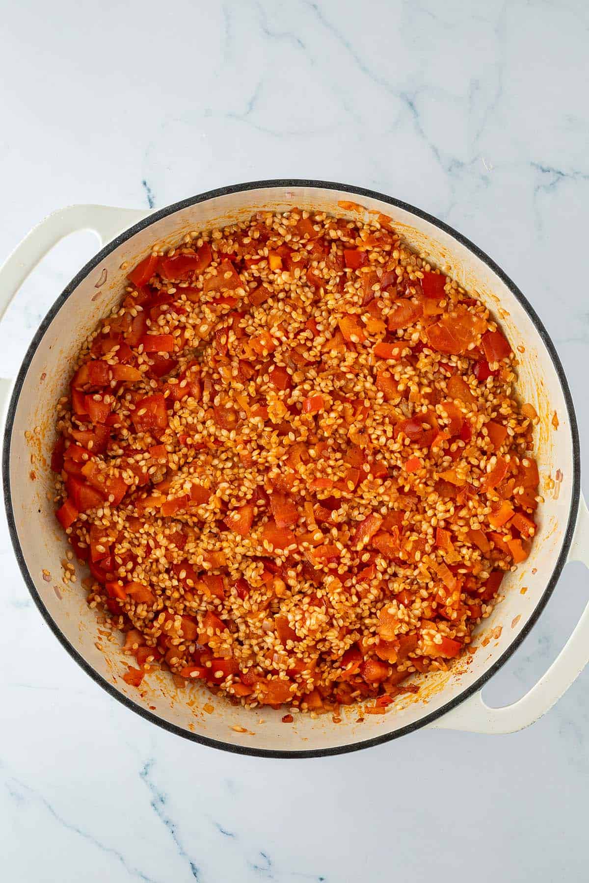 Ungekochter Paella-Reis mit angebratener Paprika, Tomaten und Zwiebeln in einer gro&szlig;en Pfanne.