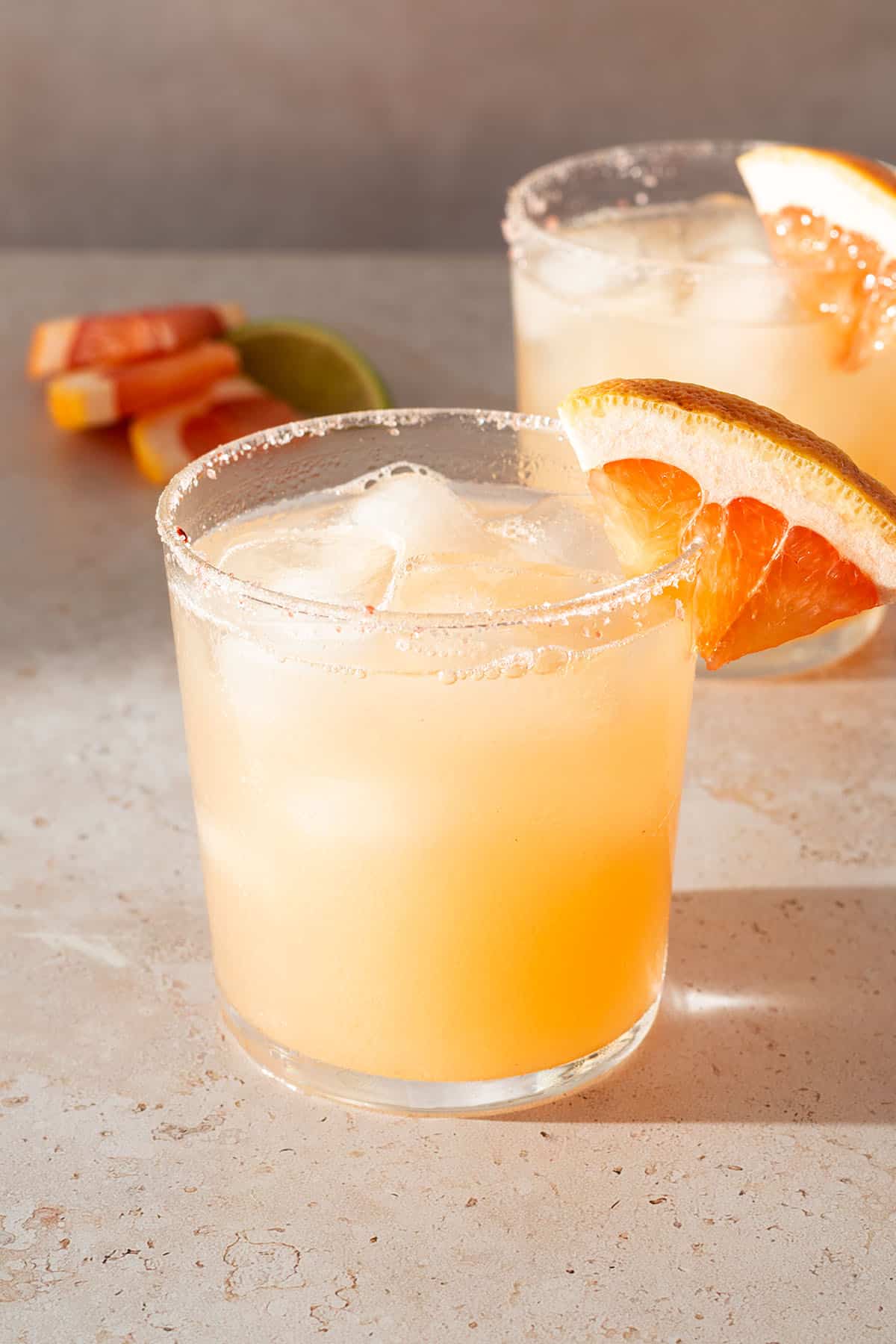 Paloma Cocktail in einem Glas mit Salzrand und einer Grapefruit-Scheibe, im Hintergrund ein zweites Glas.