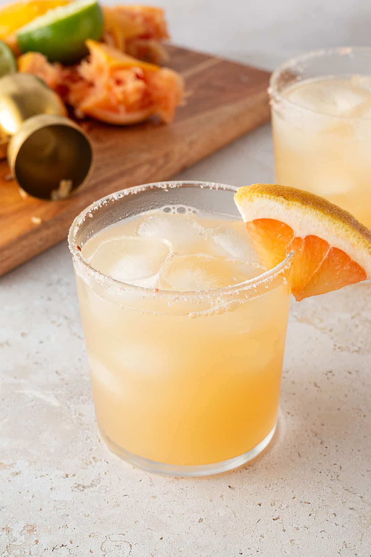 Zwei Gl&auml;ser Paloma Cocktail mit Salzrand und einer Grapefruit. Im Hintergrund ausgepresste Zitrusfr&uuml;chte.