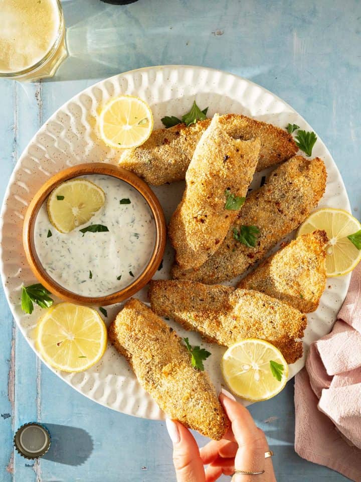 Teller mit mehreren panierten Fischfilets aus dem Airfryer, Zitronenscheiben und Kr&auml;uter-Dip, von oben fotografiert.