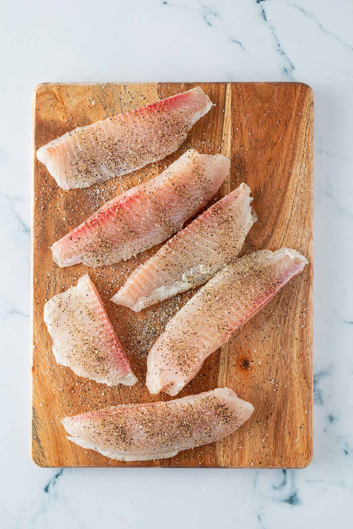 Rohe, gew&uuml;rzte Fischfilets liegen auf einem Holzbrett, bereit zum Panieren.