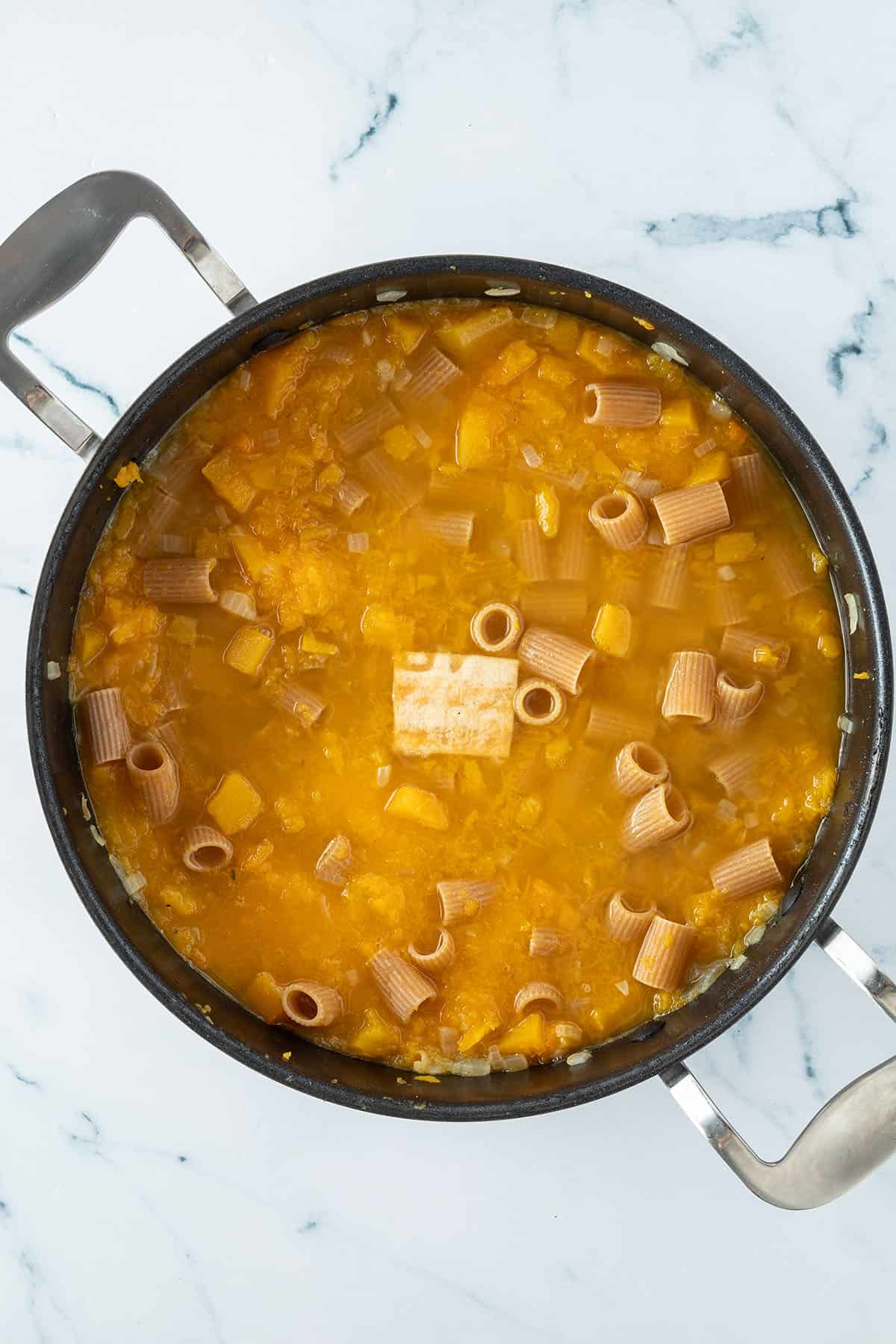 Ungekochte Nudeln und ein kleines St&uuml;ck Parmesanrinde kochen im K&uuml;rbissud in einer tiefen Pfanne.