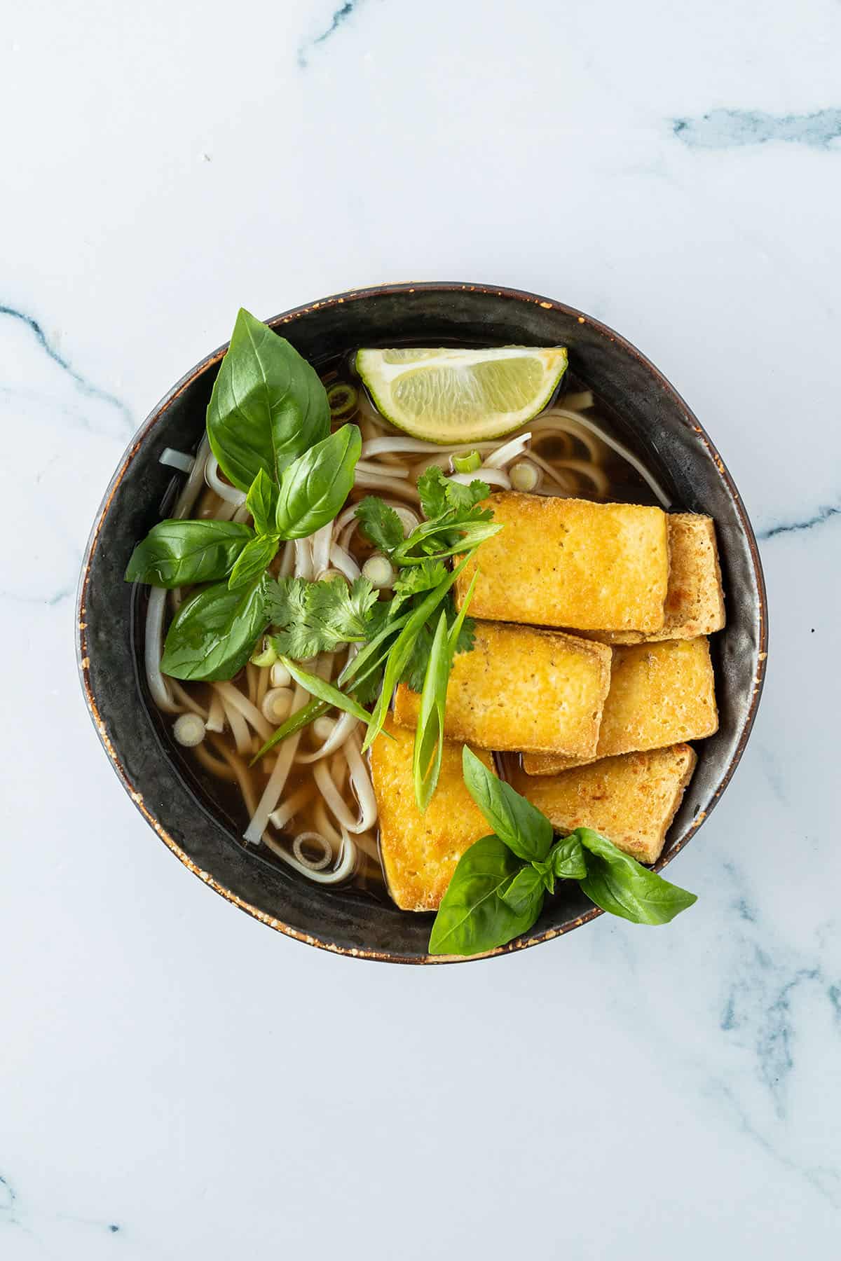 Fertig angerichtete Pho Suppe in einer Sch&uuml;ssel mit frischen Kr&auml;utern, Reisnudeln und knusprigen Tofu-Scheiben.