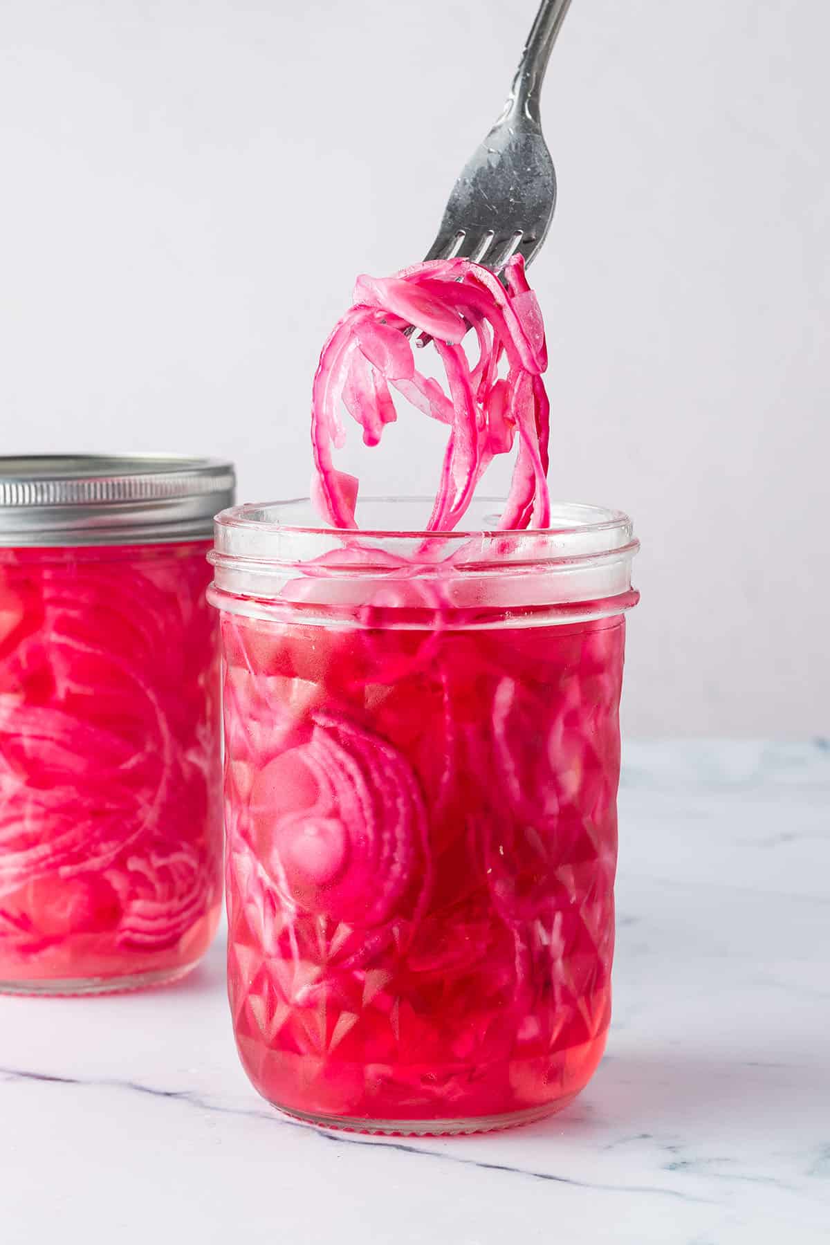 Zwei Gl&auml;ser mit Pickled Red Onions, wo eine Gabel ein paar Zwiebelringe aus einem Glas rausnimmt.