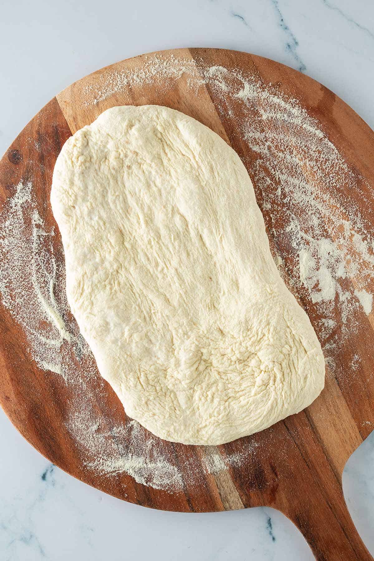 Geformter Pinsa Teig auf einem mit Semola bemehlten Holzbrett vor dem Backen.