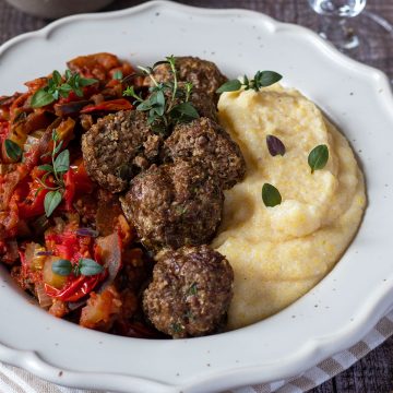 Cremige Polenta serviert mit Hackb&auml;llchen und Tomatengem&uuml;se in einem wei&szlig;en Teller, Deko im Hintergrund.