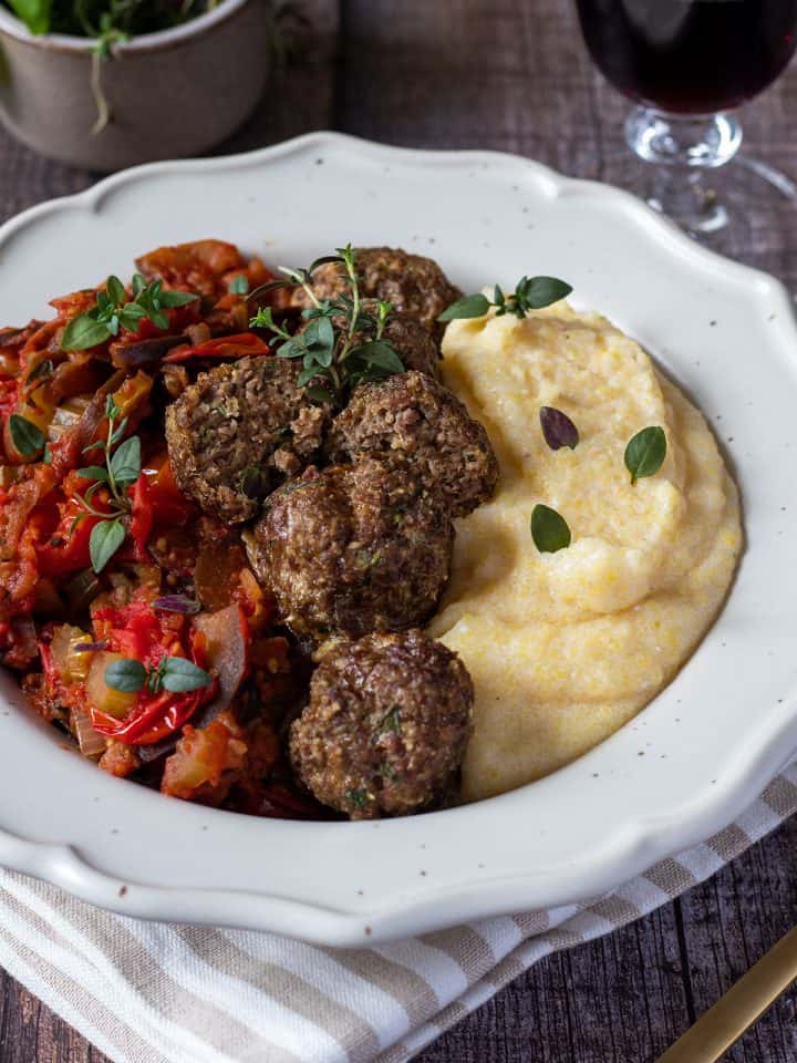 Cremige Polenta serviert mit Hackbällchen und Tomatengemüse in einem weißen Teller, Deko im Hintergrund.