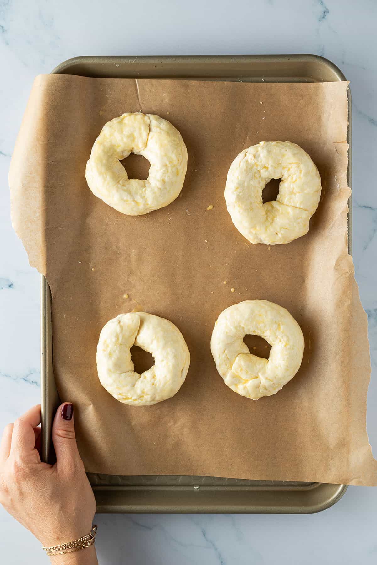 Vier rohe Bagel auf einem Blech mit Backpapier, die mit Eigelb bestrichen wurden.