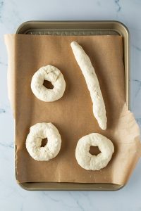 Drei rohe Teigkringel und eine l&auml;nglich geformte Teig-Wurst auf einem Blech.
