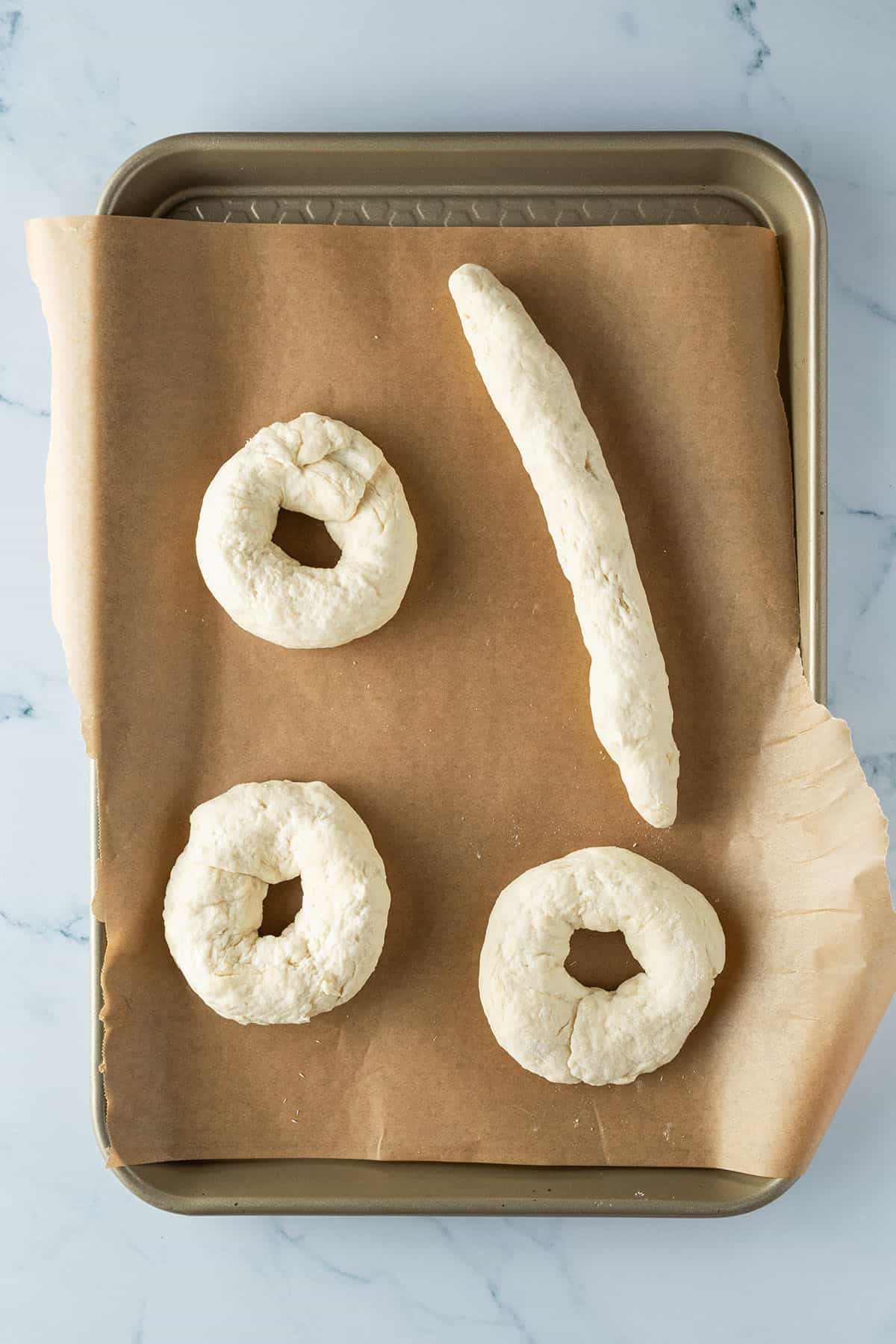 Drei rohe Teigkringel und eine l&auml;nglich geformte Teig-Wurst auf einem Blech.