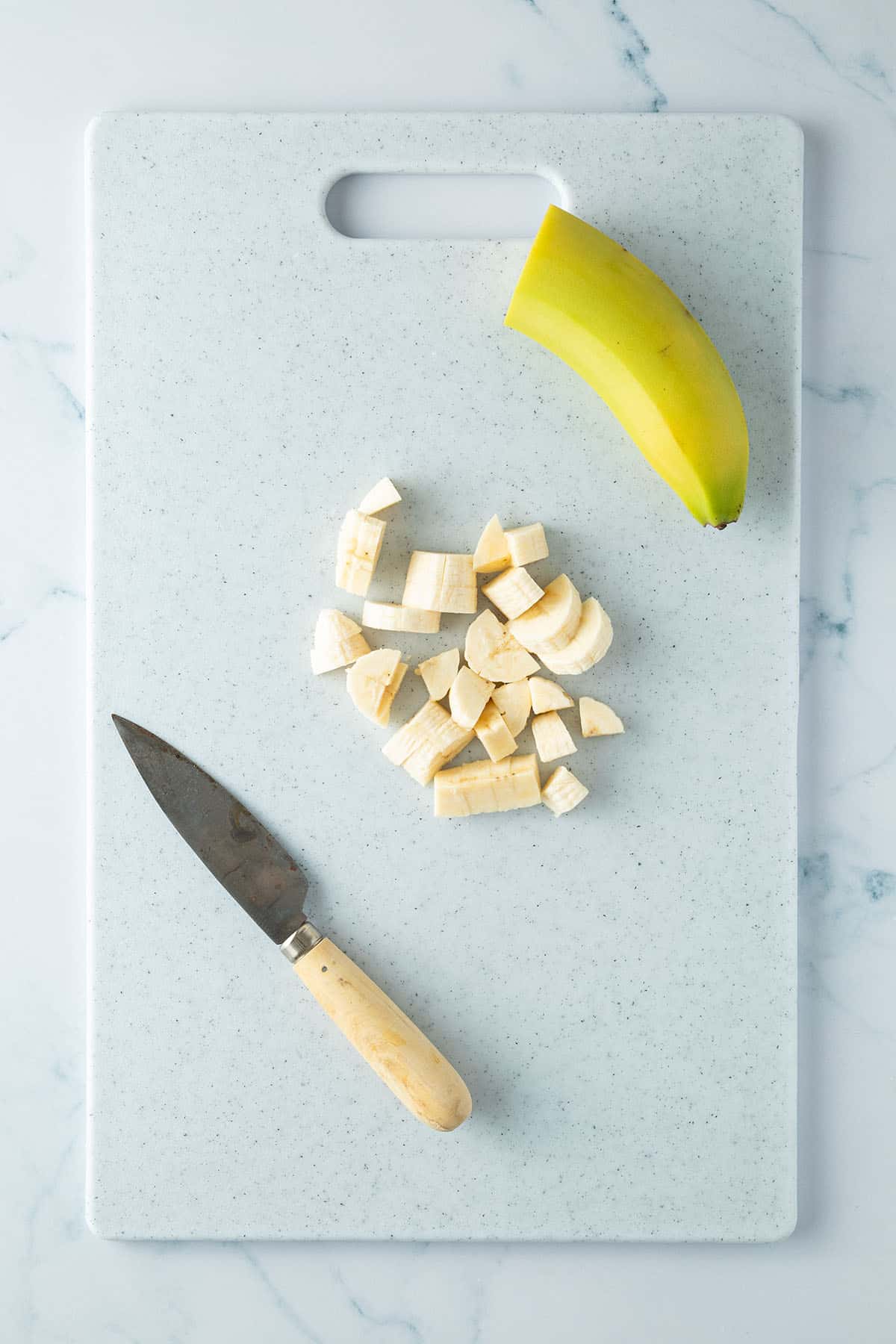 Eine halbe, kleingew&uuml;rfelte Banane auf einem Brett mit einem Messer und der zweiten Bananenh&auml;lfte.