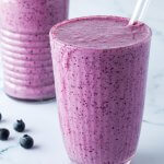 Zwei Blaubeer-Smoothies mit Strohhalmen vor einem Marmorartigen Hintergrund.