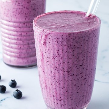 Zwei Blaubeer-Smoothies mit Strohhalmen vor einem Marmorartigen Hintergrund.