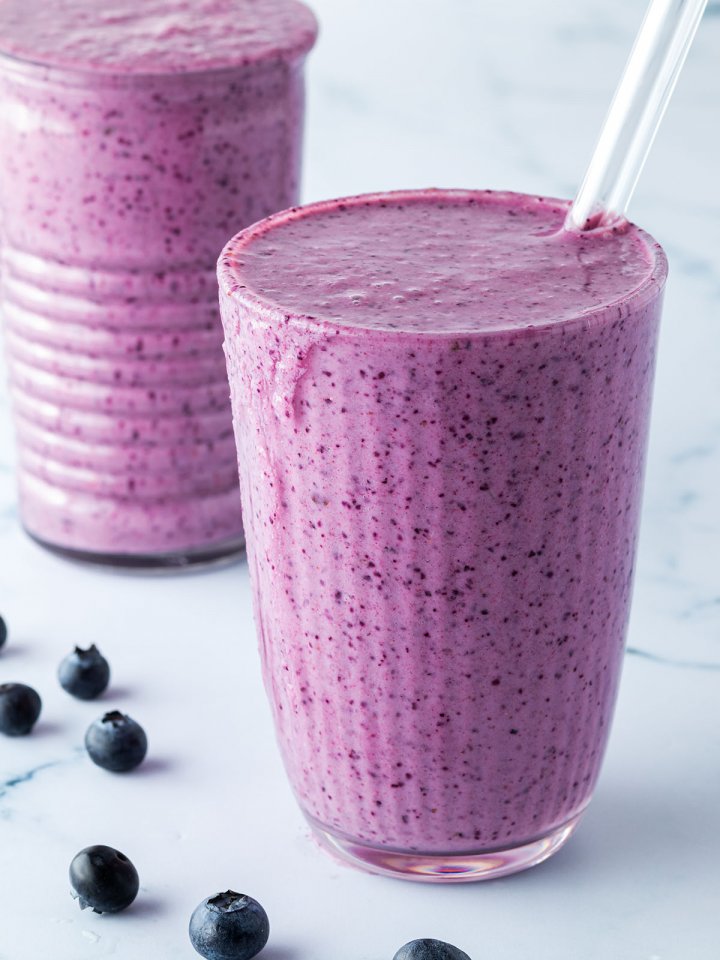 Zwei Blaubeer-Smoothies mit Strohhalmen vor einem Marmorartigen Hintergrund.
