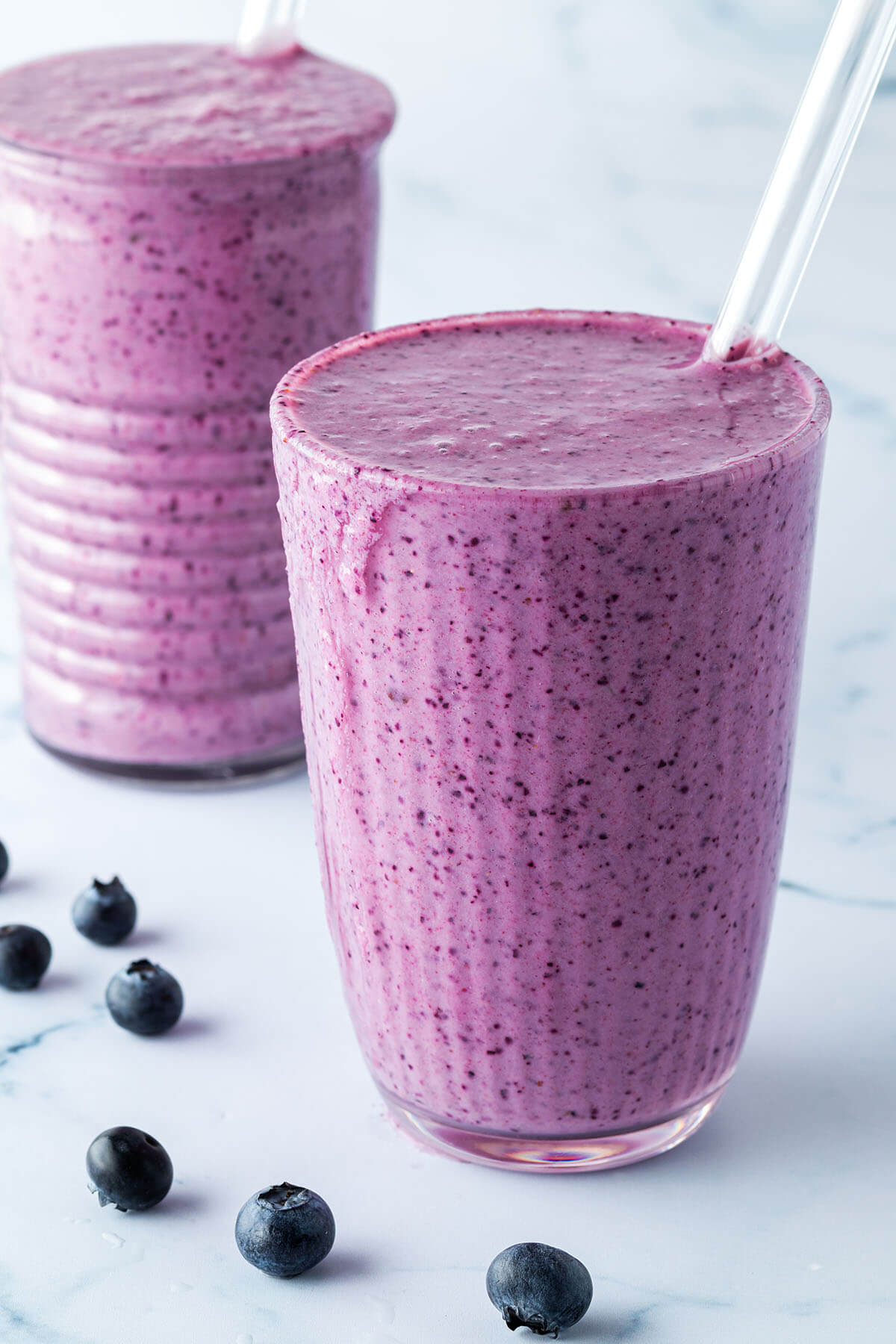 Zwei Blaubeer-Smoothies mit Strohhalmen vor einem Marmorartigen Hintergrund.