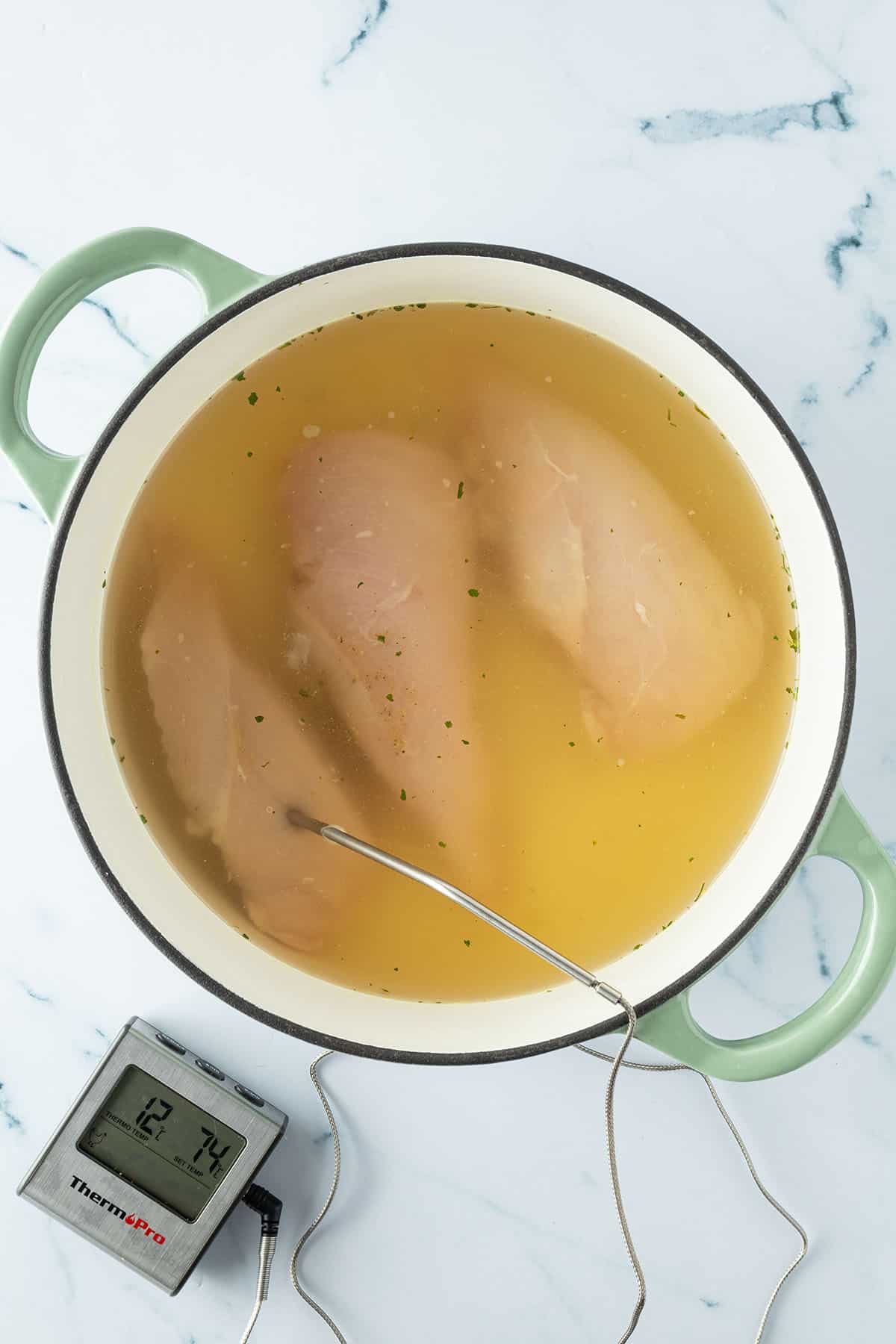 Drei H&auml;hnchenfilets in einem Topf mit H&uuml;hnerbr&uuml;he, wobei in einem der Filets ein Fleischthermometer steckt.
