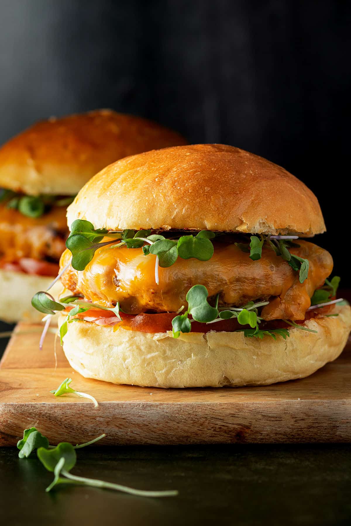 Zwei Puten Burger mit frischen Sprossen und Tomate auf einem Holzbrett vor dunklem Hintergrund.