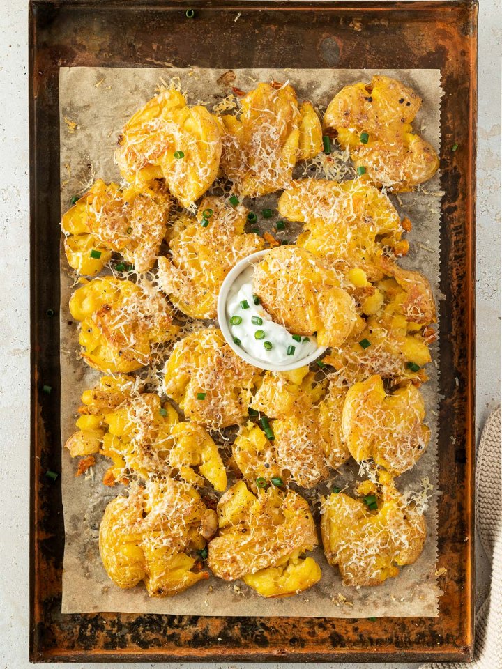 Mit Parmesan &uuml;berbackene Quetschkartoffen auf einem Blech mit einer kleinen Sch&uuml;ssel Sourcream.