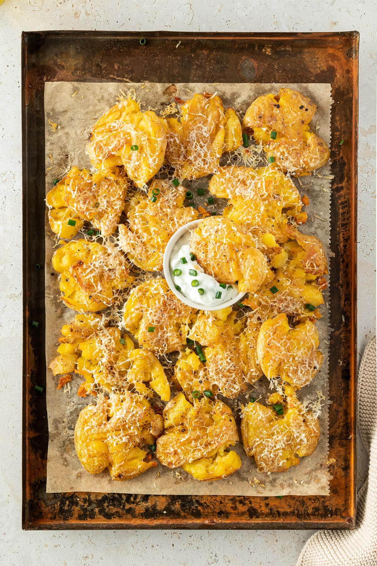 Mit Parmesan &uuml;berbackene Quetschkartoffen auf einem Blech mit einer kleinen Sch&uuml;ssel Sourcream.